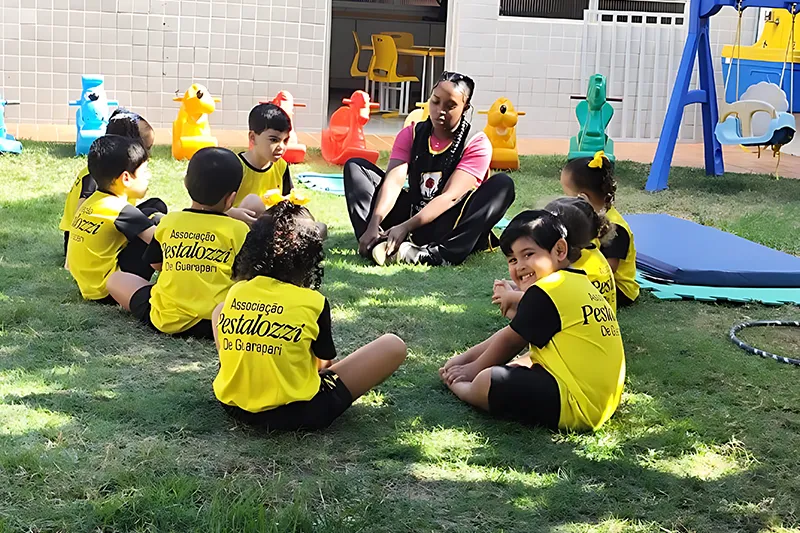 Educadora conduzindo atividade em roda com crianças no gramado do CAEE Peixinho Dourado.
